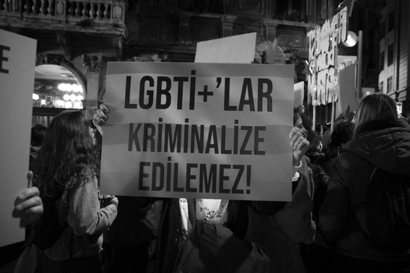 A black and white photograph taken in a protest. On the center there's a placard "LGBTI+ can't be criminalized!" written in Turkish on it.