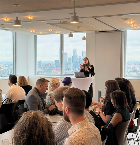 Adriana Groh addressing the breakfast reception at the German House. The New York skyline is visible through the windows behind her.