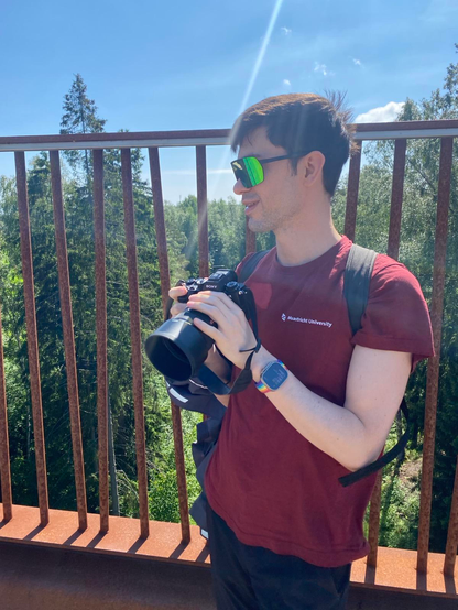 on a bright sunny day, in June 2026, here is Christopher in a red shirt, dark brown hair, white pale skin, wearing shiny green sun glasses and holding a Sony digital camera. in the background is a rusty bridge wall made out of narrow bars so that you can see through the tall evergreen trees in the background