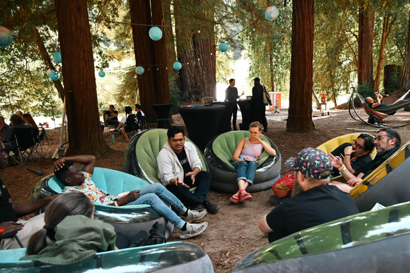 DWeb Camp attendees relaxing and conversing in inflatable lounge chairs among tall redwood trees, with teal paper lanterns strung overhead and a hammock visible in the background.