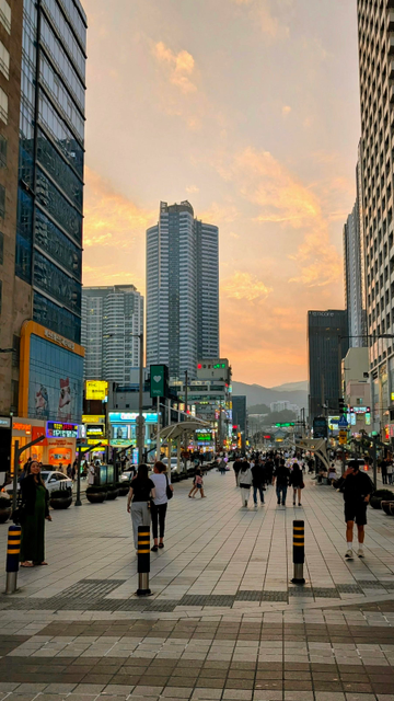 Colour photo of a main street in Haeundae, Busan. We're looking towards the mountains here. The sky has a mixture of pastel colours whilst on the streets, modern high rise buildings and bright lights dominate. People are enjoying an evening stroll out of the searing sunshine.
