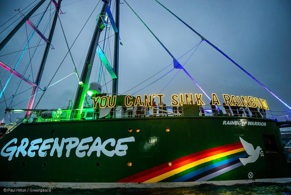 Das Greenpeace Schiff Rainbow Warrior. Auf dem grünen Rumpf steht in großen, gut lesbaren weißen Buchstaben der Schriftzug "GREENPEACE", daneben ein leuchtender Regenbogen mit einer weißen Friedenstaube, die einen Ölzweig hält. Entlang des beleuchteten Oberdecks, oberhalb des Rumpfes, sind große, gelbe Neonbuchstaben angebracht, die die Botschaft formen: "YOU CAN'T SINK A RAINBOW". 