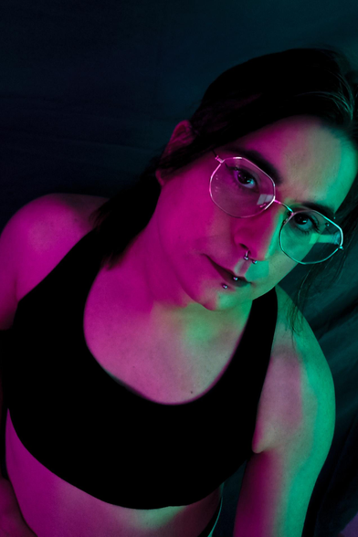 a white fem taken from an high angle, standing in bra, looking to the camera, she wears octagonal glasses and has piercings to the ear, eyebrow, septum and lower lip.
she has a soft nude make up with dark lips and liner tails and her face is illuminated by a dark magenta light on one side and and light cyan on the other.
her long hair are tied letting hang just to strand that frame her face