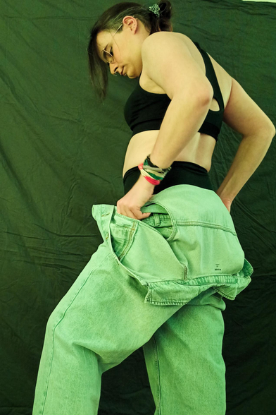 under a yellowish light, Melo is undressing, their sleeveless denim overall is down their waist