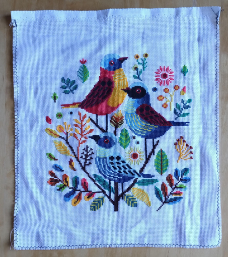 Finished cross stitch pattern with three colourful birds in the middle, surrounded by leaves.