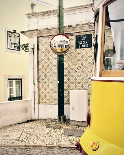 Yellow tram rounding a corner beside a tiled building façade in a narrow street. A black street sign reads “Rua das Escolas Gerais”, while a convex traffic mirror reflects the tram from another angle. Traditional paving stones, a decorative wall pattern and an old-style street lamp complete the Lisbon streetscape.