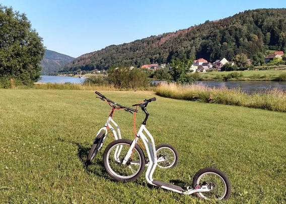 Zwei weiße Tretroller stehen auf einer grünen Wiese direkt an der Elbe. Im Hintergrund fließt der Fluss vorbei, dahinter liegt ein kleines Dorf mit roten Dächern am bewaldeten Hang der Sächsischen Schweiz. Der Himmel ist strahlend blau und vermittelt eine ruhige, sommerliche Stimmung.