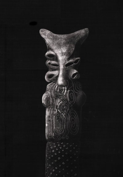 A black and white scratchboard drawing of a vertical anthropomorphic totem or fetish figure, characterized by intricate textures. The top of the figure is anvil-shaped, positioned above a mask-like face with concave eye sockets and a central nose/mouth structure. The mid-section features a dense pattern of intertwined spirals and figure-eight motifs, while the base displays a geometric grid of textured diamonds and hexagons. The entire piece is rendered against a deep, dark black background.