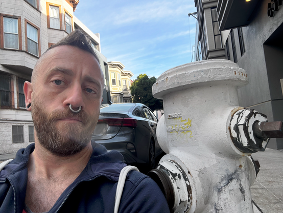 Bearded man with a septum ring, undercut, and blue eyes gives deadpan in a blue hoodie beside a weathered white fire hydrant. Foggy SF street. We are both structurally load-bearing.