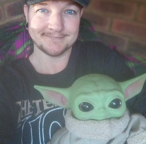 White trans man(me) with dark facial hair and piercing blue eyes. In smiling at the camera. I sit in a chair, and I'm wearing a grey tee shirt. On my lap is a Baby Yoda/Grogu doll.
