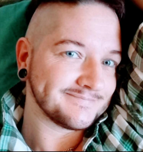 Trans man (me) with dark hair and facial hair. He has piercing blue eyes, and smiles at the camera with a slightly suggestive look. He rests on a blue pillow with one arm behind his head, and wears a grey green and white plaid shirt.