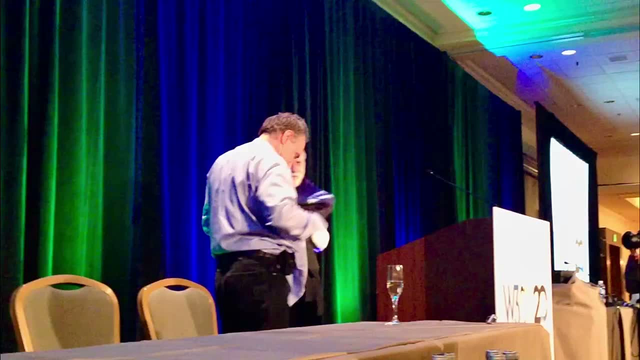 Two men stand on stage. One removes his shirt and reveals a T-shirt with text in the front "I didn't invent the Internet." The other man dons over his white shirt a t-shirt that reads on the front "I invented the Internet." They smile in the lighthearted moment. A crowd gathers at the foot of the stage as people laugh and film.