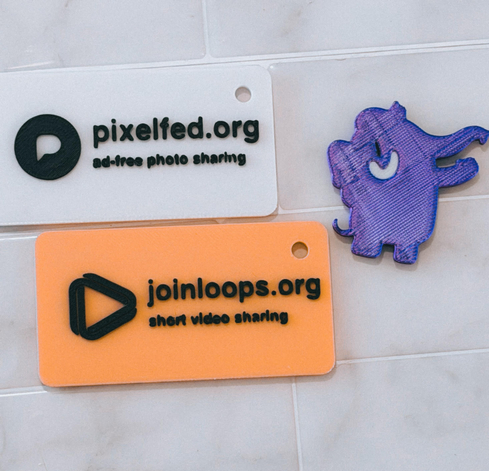 An image of a 3d printed white card with black text with the Pixelfed logo and pixelfed.org in large text, with ad-free photo sharing underneath, below that is a yellow background card with the loops logo and joinloop.org in large text with short video sharing underneath. To the right is a 3d-printed mastodon mascot mastodon