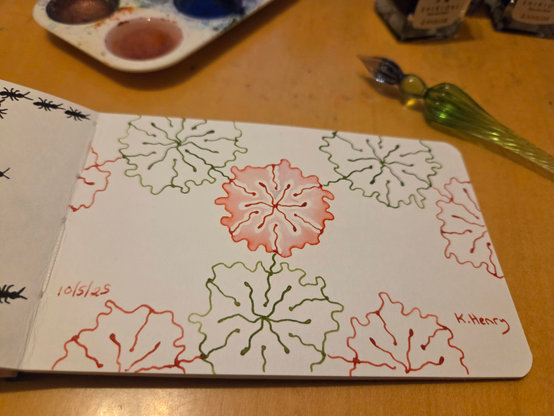 Hand drawn generative/procedural art in ink on an open page of my sketchbook. The abstract pattern looks a bit like a network diagram with flowers at the nodes. Next to my sketchbook is my glass dipping pen and some ink bottles.