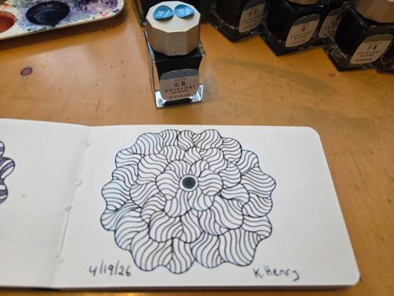 Hand drawn generative/procedural art in ink on an open page of my sketchbook. The abstract pattern is floral. The ink I drew with is in the bottle next to my sketchbook.