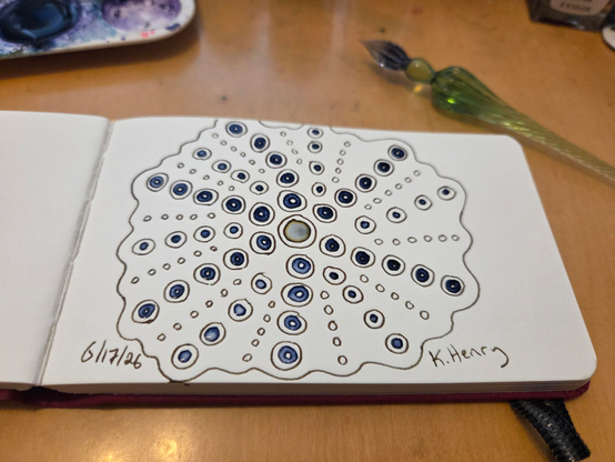Hand drawn generative/procedural art in ink on an open page of my sketchbook. The abstract pattern looks a bit like a sea urchin. My glass dipping pen is next to my sketchbook.