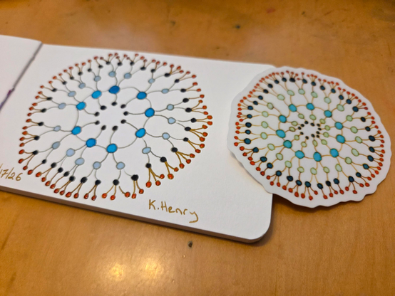 Hand drawn generative/procedural art in ink on an open page of my sketchbook. The  abstract pattern looks kinda like a beaded necklace. A sticker, made from a similar drawing, is next to my sketchbook.