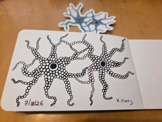 Hand drawn generative/procedural art in ink on an open page of my sketchbook. The abstract pattern looks a bit like two interconnected nerve cells. A sticker, with a similar pattern, is next to my sketchbook.