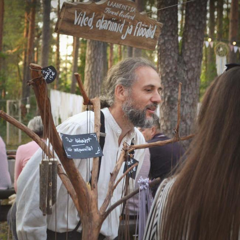 In a pine forest, a bearded man is standing behind a display made from a branch, on which whistles are hung. He is smiling slightly at someone outside the frame of the photograph.