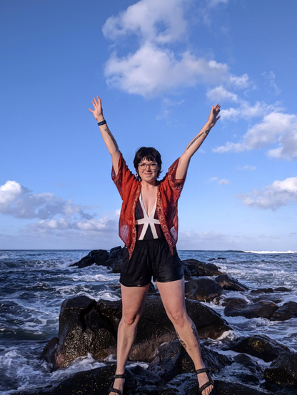 A photo of me standing on the rocky coast of a beach on Oahu Island in Hawaii. I'm standing on large black igneous rocks, with more large rocks behind me. It's a beautiful sunny day, with a few small clouds in the sky. I'm wearing a bathing suit and black shorts with a red kimono style wrap and sandals. My arms are high up in the air! I took this photo as the tide was coming in, and it took multiple tries as me and my camera kept getting splashed with large waves!