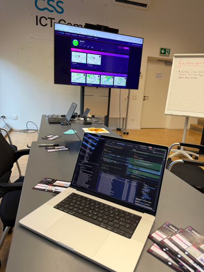 A meeting room at the CSS ICT Campus during a Hackergarten evening. A large wall-mounted screen shows a FitPub user profile with recent peaks and a grid of running activity maps. Two laptops sit on grey tables, the nearer one open with a code editor full of colourful syntax highlighting. FitPub flyers are spread across the tables next to a marker pen and a bowl of crisps. On the right a flip chart headed Hackergarten lists the evening's projects: FitPub in Java and HTML, a LinkedIn fact check plugin, and an Arduino motor controller."
