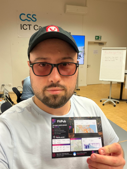 A selfie in the same room. A man in a white t-shirt, glasses and a dark cap with the red Vivaldi logo holds up a FitPub flyer towards the camera. Behind him are the CSS ICT Campus sign, a screen, and the flip chart listing the evening's projects with vote counts beside them.