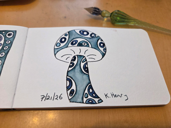 Hand drawn generative/procedural art in ink on an open page of my sketchbook. The abstract pattern looks a bit like a mushroom with spots on the stem and cap. My glass dipping pen is next to my sketchbook.