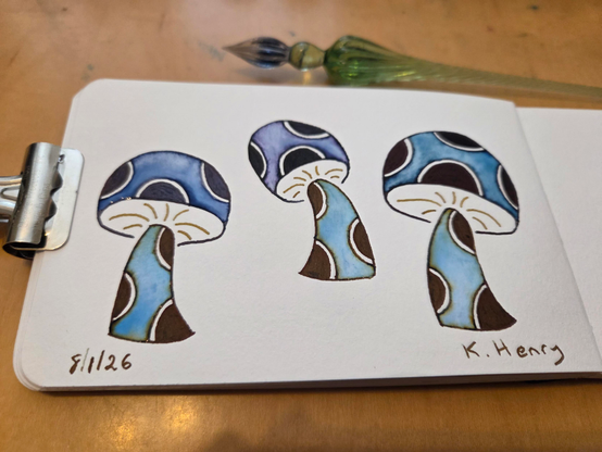 Hand drawn generative/procedural art in ink on an open page of my sketchbook. The abstract pattern has three abstract mushrooms with polka dots on the stems and caps. My glass dipping pen is next to my sketchbook, which is held open with a binder clip.