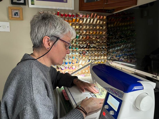 A photograph of me (lucy) sitting at my sewing machine with a wall display of coloured reels of cotton behind me