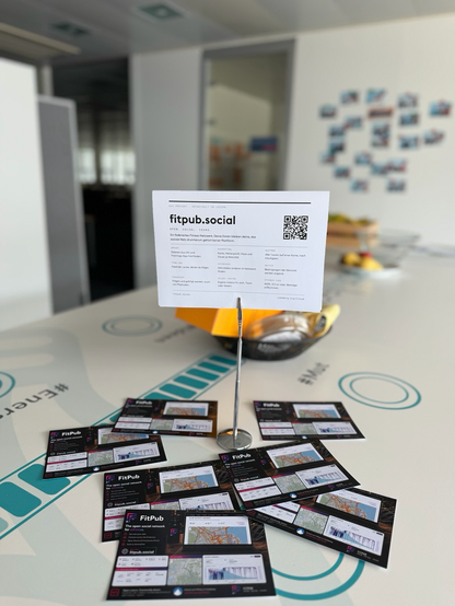 Photo of a white table in a modern office. A metal stand holds a white printed card headed “DAS PROJEKT · ENTWICKELT IN LUZERN”, large “fitpub.social”, the line “OPEN. SOCIAL. YOURS.”, and a German paragraph describing a federated fitness network. The card lists features in two columns (IMPORT, TIMELINE, FOLLOWING / AUSWERTUNG, ENTDECKEN, SELF HOST / HEATMAP, WETTER, OFFENER CODE) plus a QR code and codeberg.org/fitpub. Around the base of the stand lie roughly nine dark business cards showing the FitPub logo, the English phrase “The open social network for athletes”, screenshots of maps and activity graphs, and fitpub.social. Behind the stand is a black wire basket with yellow packets and bananas. The table surface has large teal graphics including “#Energies” and “#Mist”. Glass doors and a wall of small photos appear in the background.