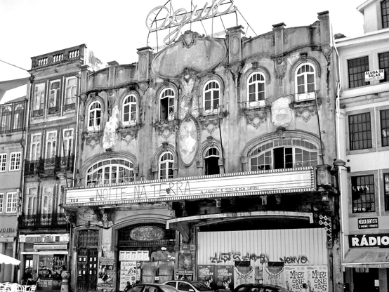 A imagem mostra a fachada histórica e em ruínas do antigo cinema Águia d'Ouro, na Praça da Batalha, no Porto, Portugal. História: Este cinema de longa tradição encerrou as suas portas no final de 1989 e, desde então, tem estado abandonado, o que levou a um forte estado de degradação do edifício ao longo dos anos. Arquitetura: A fachada, de estilo Art Nouveau em declínio/ecletismo clássico, ainda exibe a inscrição marcante «Águia» no telhado, bem como antigos cartazes de cinema e inscrições.