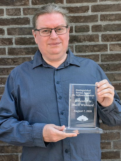 Mark J. Wielaard (mjw) holds the desk-standing plaque of his award.  The award is clear lucite and reads: “Distinguished Service award for Software Freedom / presented to Mark Wielaard / August 7, 2026”.  The award also has SFC's tree logo etched below the text.