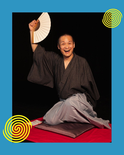 A rakugo performer in kimono, Kanariya Eishi, is sitting on a Japanese cushion. He is holding up an open Japanese traditional fan above his head with his right hand.