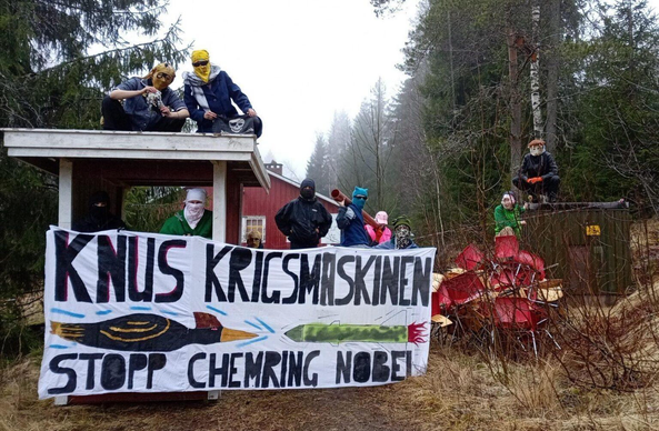 A group photo of 8 of the people squatting the house. Everyone is masked to conceal their identity. In front there is a large banner with the text "knus krigsmaskinen, stopp Chemring Nobel". On the banner is an illustration of a bird local to the area, flying beak first against a missile, as a symbol of nature's resistance against war. In the background the squatted house is visible. There are trees and nature around.