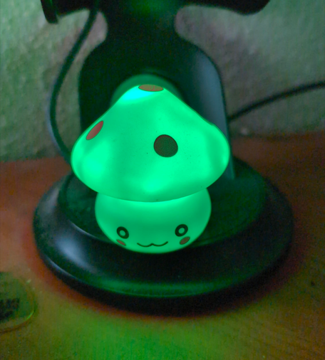 A close up picture of a battery powered nylon mushroom which contains a color changing LED inside of it. The mushroom is illuminated in green, and has has a cute smile, blushing cheeks and 2 round eyes. 