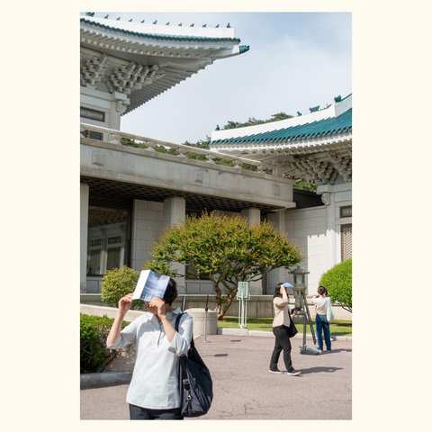 Two people holding a brochure in front of their faces standing outside a traditional building with tiled roofs and ornamental eaves