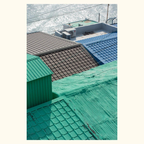 Overlapping rooftops with different materials and colors including corrugated metal and tiles near a body of water