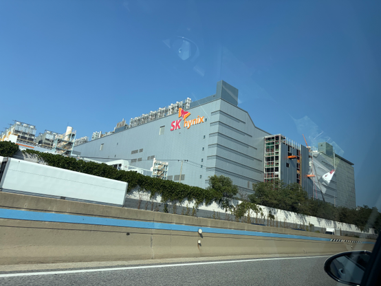Photo taken from inside a car on a highway, looking out over a concrete median toward a large gray industrial facility with an "SK hynix" logo on its upper facade. A section of the building under construction with scaffolding is visible on the right, alongside cooling towers and machinery. A truck trailer and roadside greenery are in the foreground under a clear blue sky.