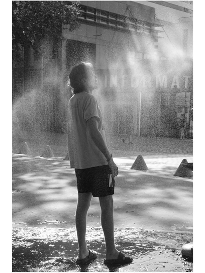 photo en noir et blanc d'un homme de dos qui profite des brumisateurs. La lumière du soleil fait briller les gouttelettes et donne un aspect un peu magique.